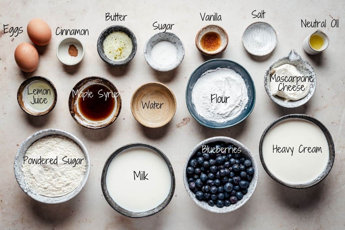 Ingredients for blueberry crepes in bowls displayed on a light surface, including eggs, cinnamon, butter, sugar, vanilla, salt, neutral oil, lemon juice, maple syrup, water, flour, mascarpone cheese, powdered sugar, milk, blueberries, and heavy cream.
