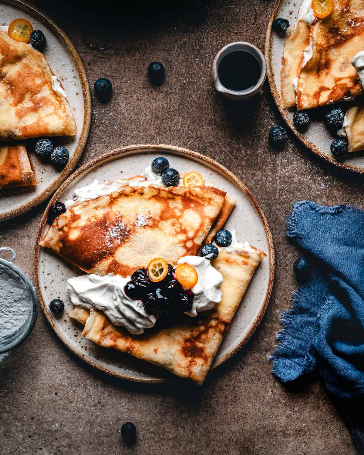 A plate of blueberry crepes topped with whipped cream, blueberry compote, blueberries, and sliced kumquats, surrounded by more plates of crepes, blueberries, a blue napkin, and a cup of syrup on a dark surface.