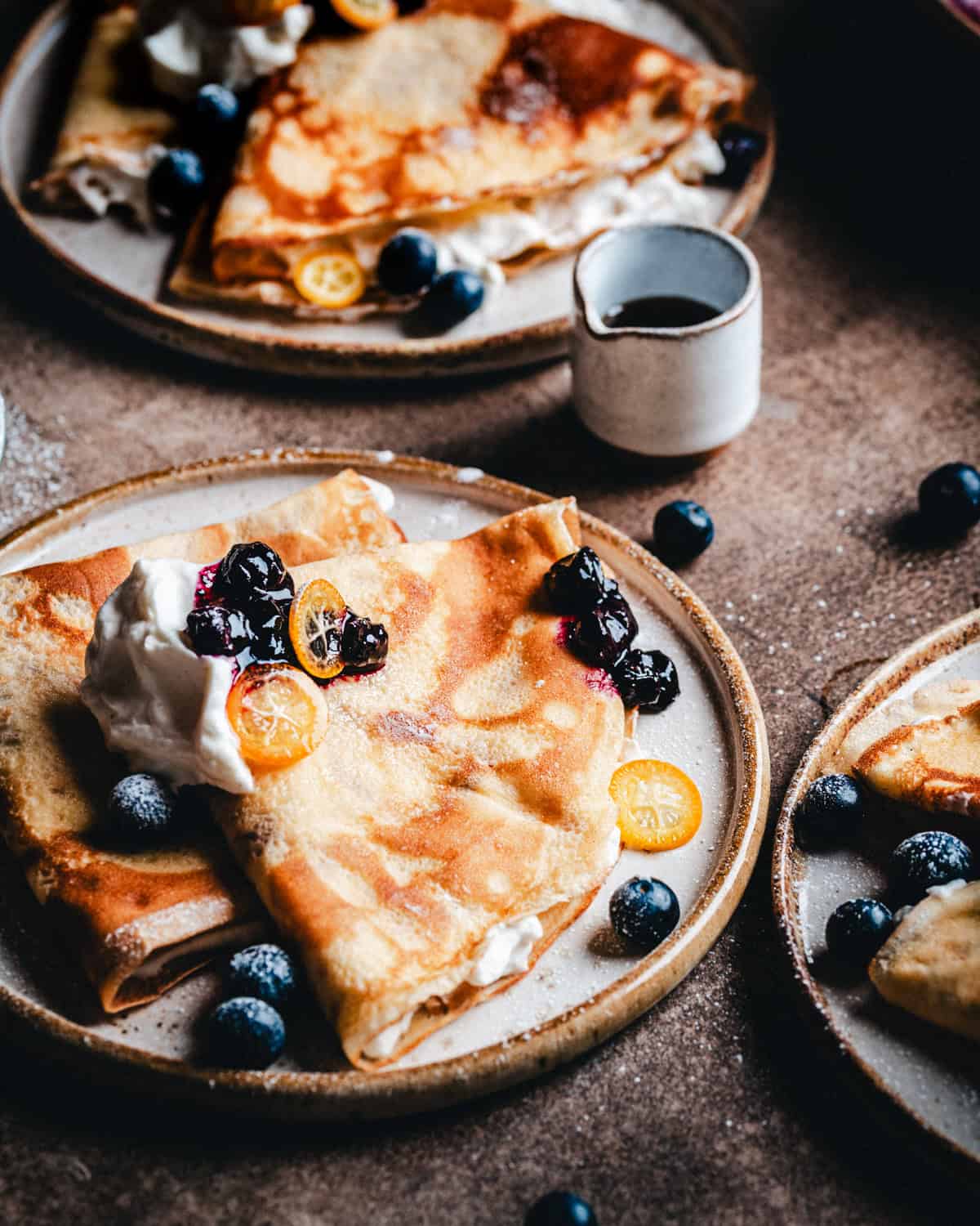 Plates of crepes topped with whipped cream, blueberries, and sliced kumquats, with a small cup of syrup nearby on a rustic surface.