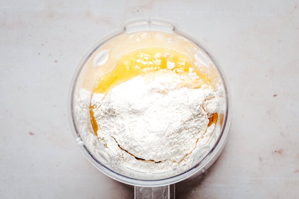 A blender bowl filled with flour, melted butter, eggs, and milk ready to be mixed, viewed from above on a light countertop.