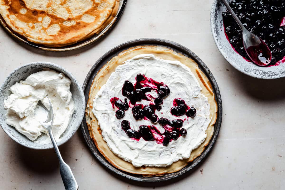 A crepe on a plate topped with whipped cream and blueberry compote, surrounded by bowls of more whipped cream, blueberry compote, and a stack of plain crepes on a light surface.