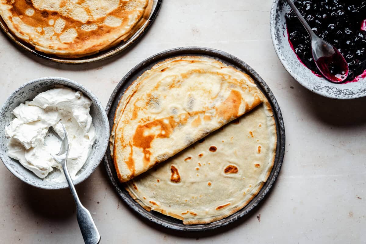 A plate with two thin crepes, one folded in half, is surrounded by a bowl of whipped cream with a spoon and a bowl of blueberry compote with a spoon on a light countertop.