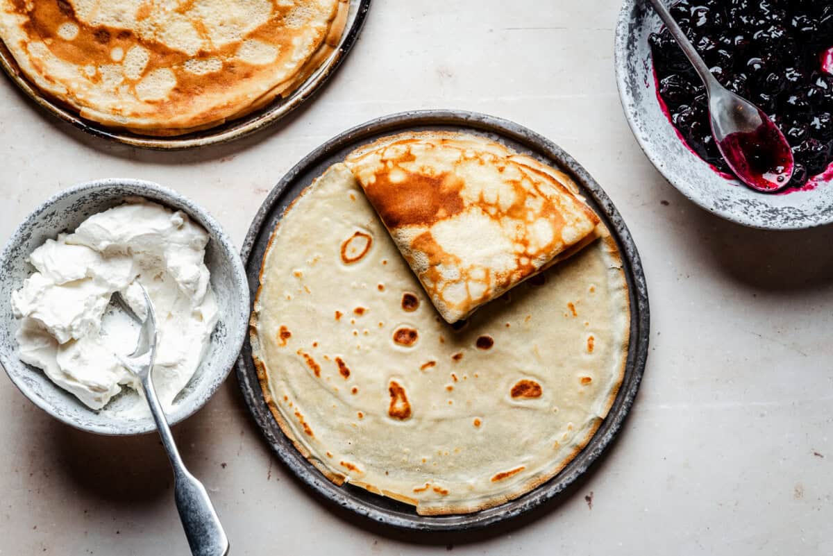 A plate of thin crepes, one folded, is shown with bowls of whipped cream and blueberry compote on the side. A fork rests in the bowl of cream, all set on a light surface.
