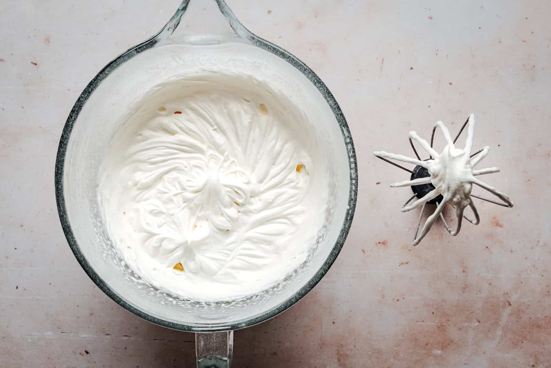 A glass bowl filled with whipped cream sits on a light surface. Next to it lies a metal mixer whisk coated in the whipped cream.