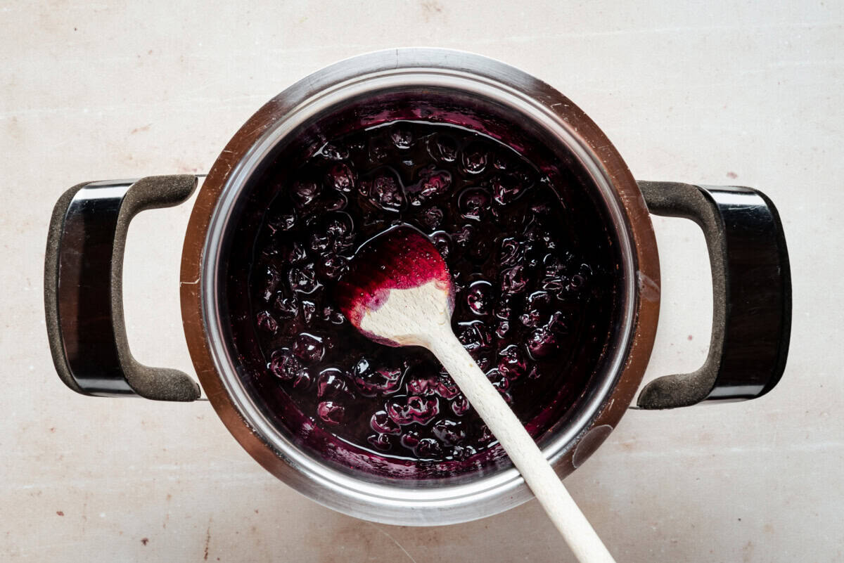 A stainless steel pot filled with dark purple blueberry jam being stirred by a wooden spoon, viewed from above on a light-colored surface.