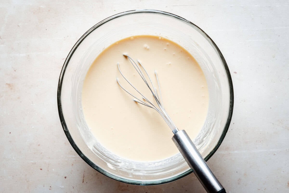 A glass bowl filled with smooth, pale batter, with a metal whisk resting inside, sits on a light-colored surface.