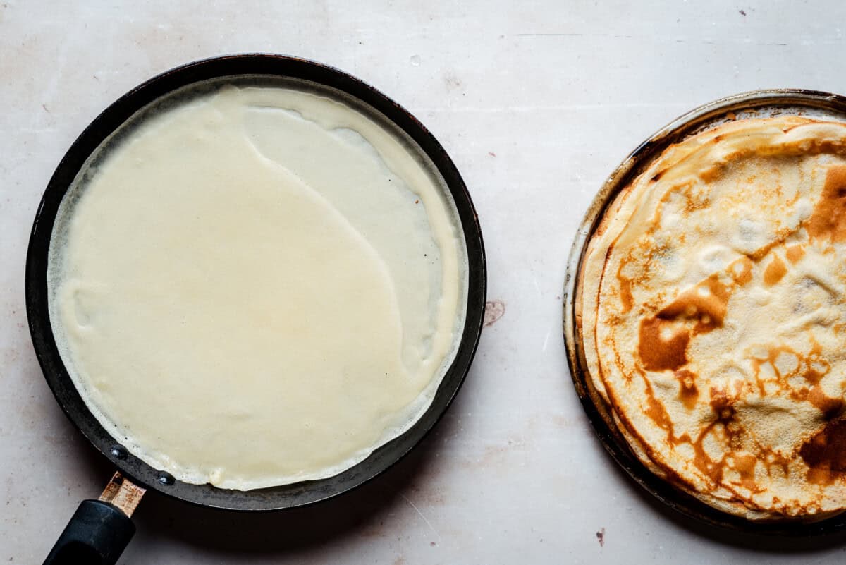 A crepe cooking in a skillet next to a plate stacked with cooked crepes on a light-colored surface.