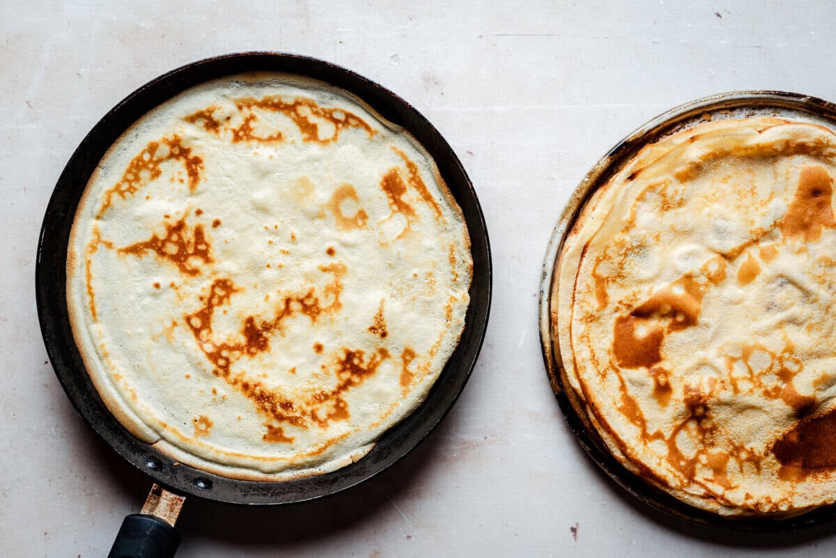 A frying pan with a cooked crepe sits next to a plate stacked with more golden-brown crepes on a light-colored surface.