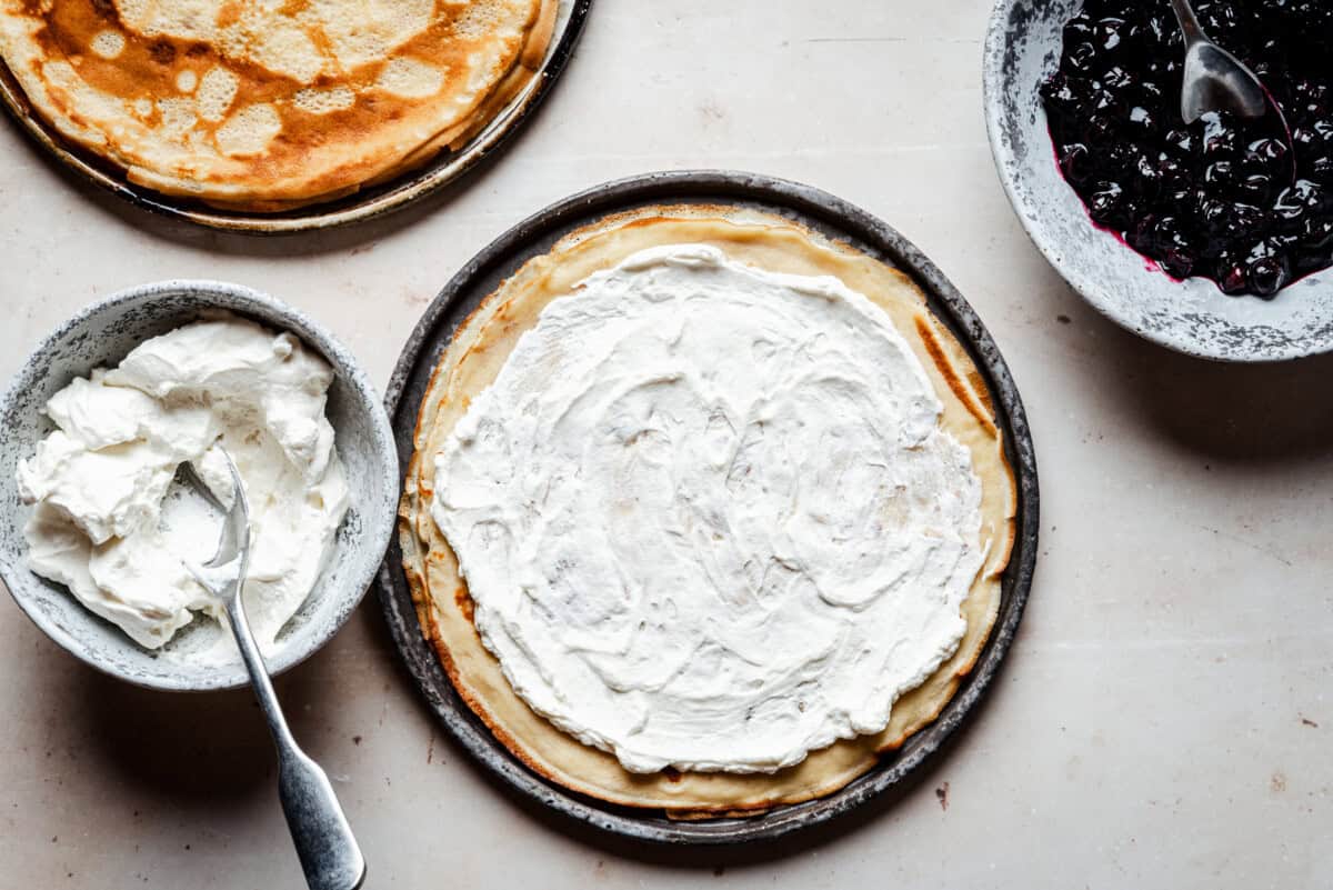 A plate with a crepe spread with whipped cream sits beside bowls of whipped cream, blueberry sauce with a spoon, and a stack of plain crepes on a light surface.