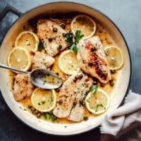 A skillet with cod piccata garnished with lemon slices, capers, and fresh parsley; a spoon rests on the side, and a white napkin is placed nearby on a dark surface.