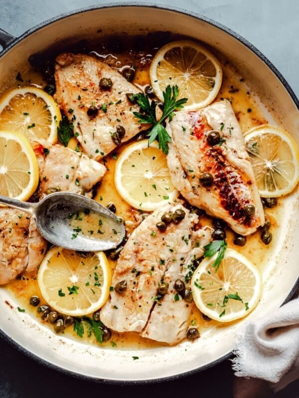 A skillet with cod piccata garnished with lemon slices, capers, and fresh parsley; a spoon rests on the side, and a white napkin is placed nearby on a dark surface.