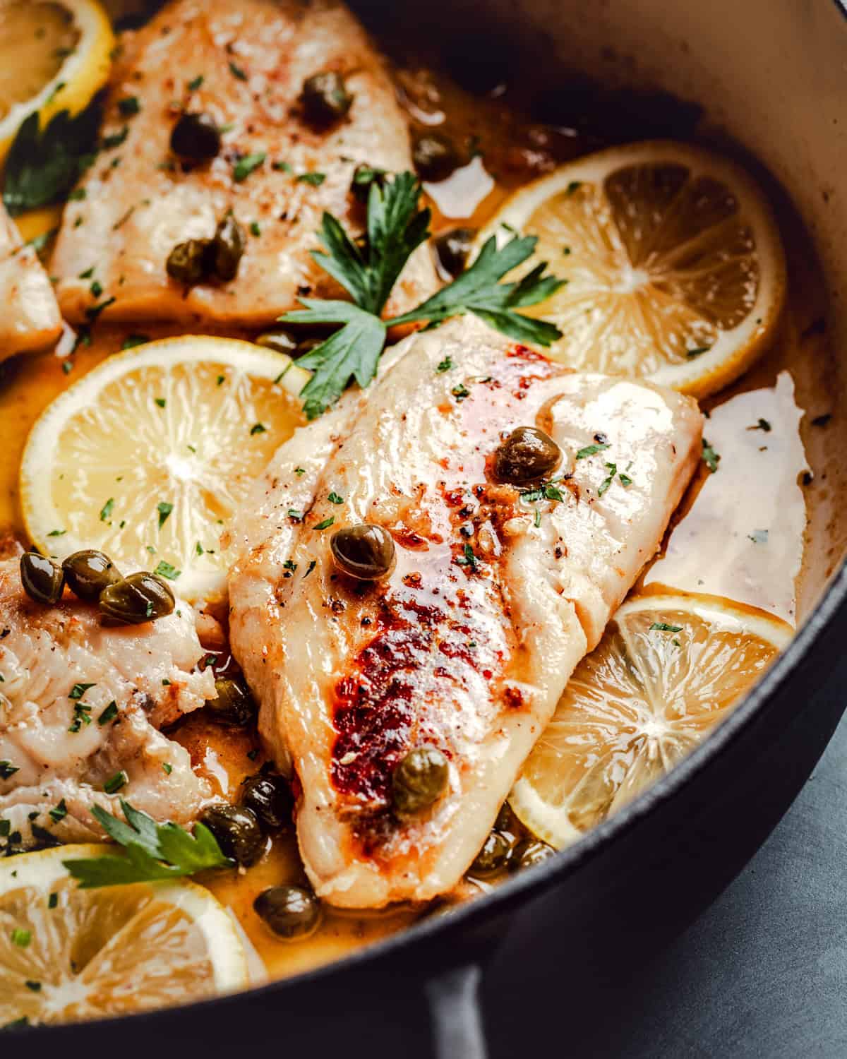 Pan-seared cod fillets garnished with capers and parsley, served with lemon slices in a flavorful piccata sauce inside a skillet.