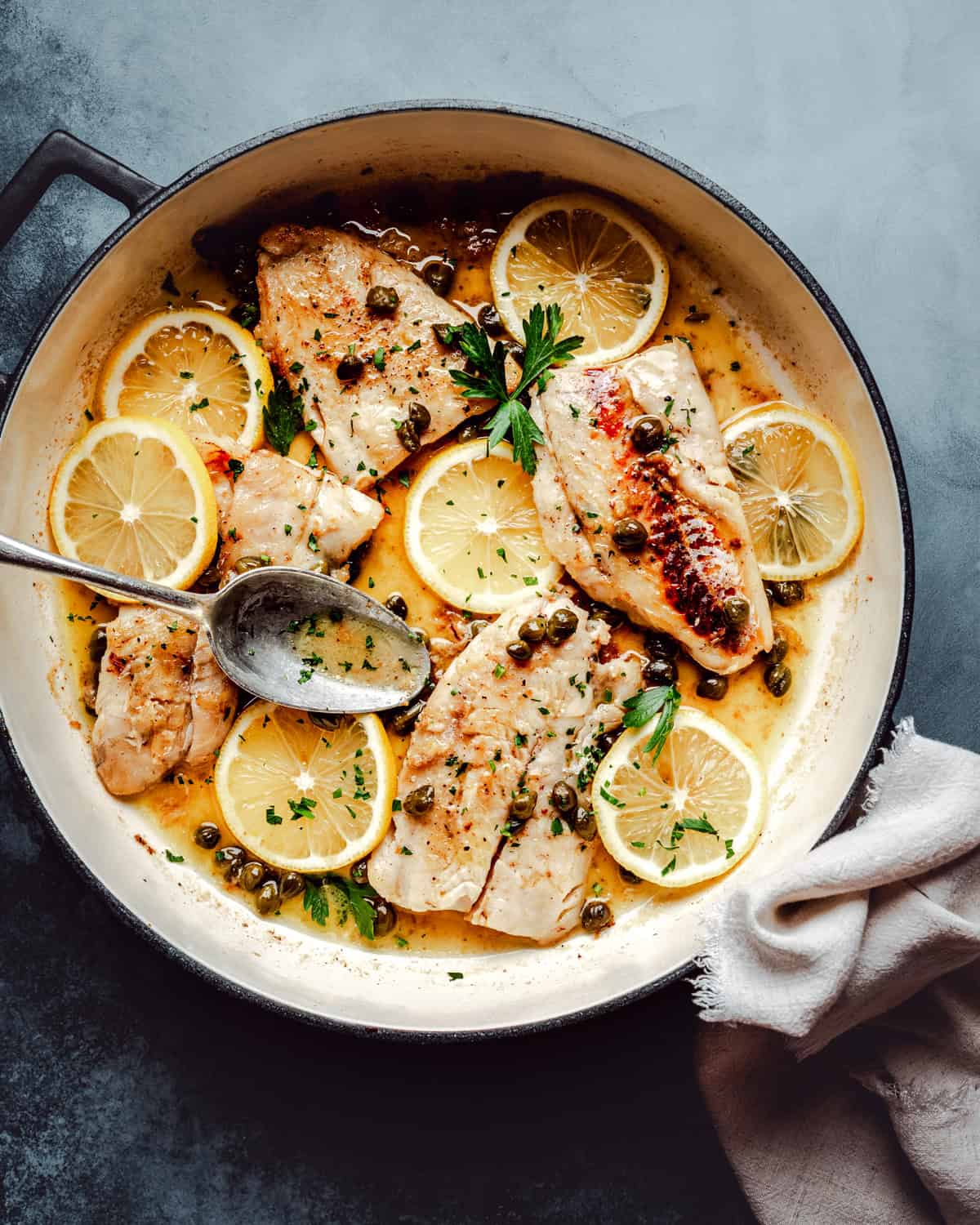 A skillet filled with cooked cod fillets topped with lemon slices, capers, and fresh parsley, resting in a light, piccata sauce. A spoon is poised over the fish, and a white cloth sits nearby.