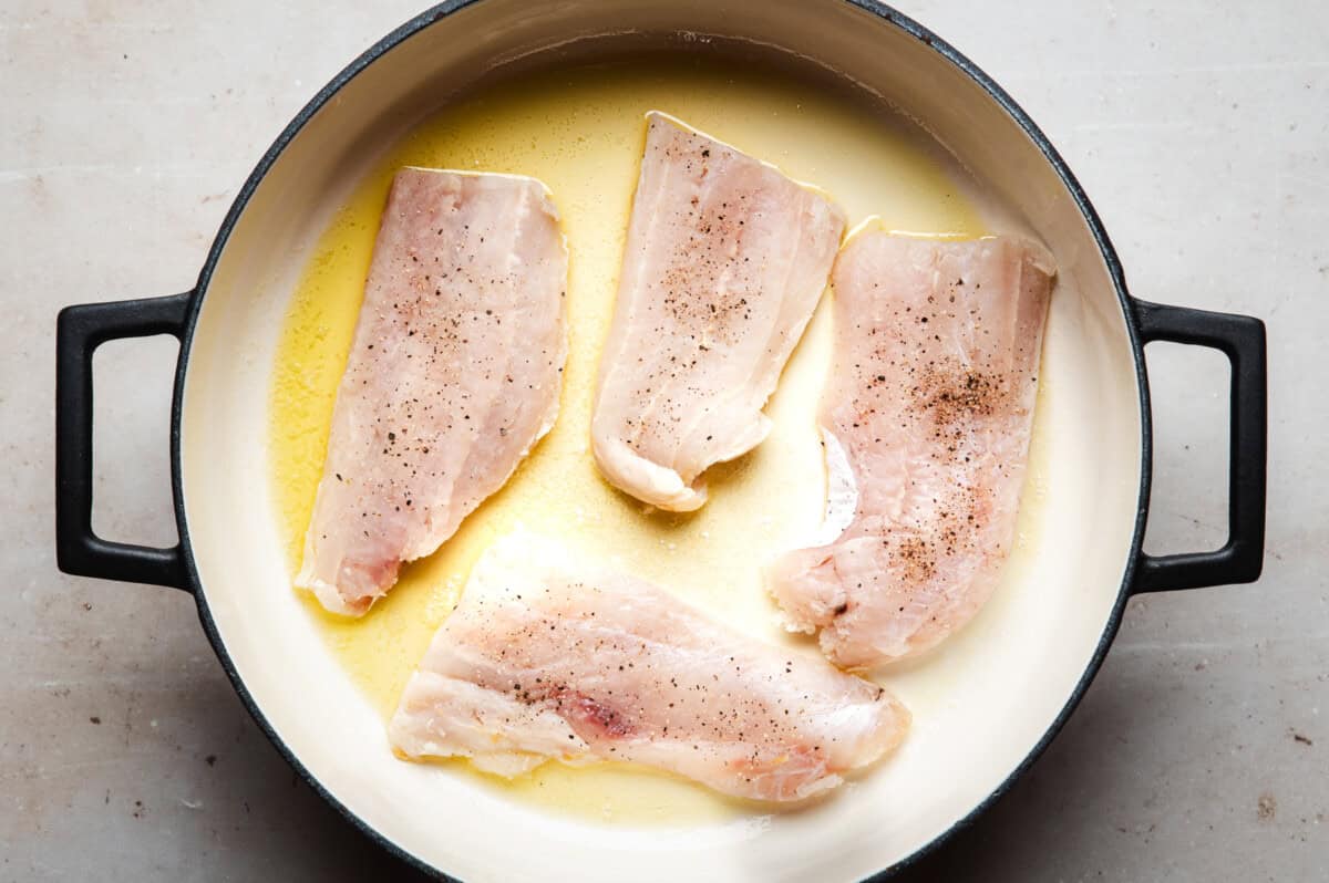 Four raw cod fillets seasoned with salt and pepper are arranged in a round, white enameled pan with melted butter or oil. The pan has black handles and sits on a light-colored surface.