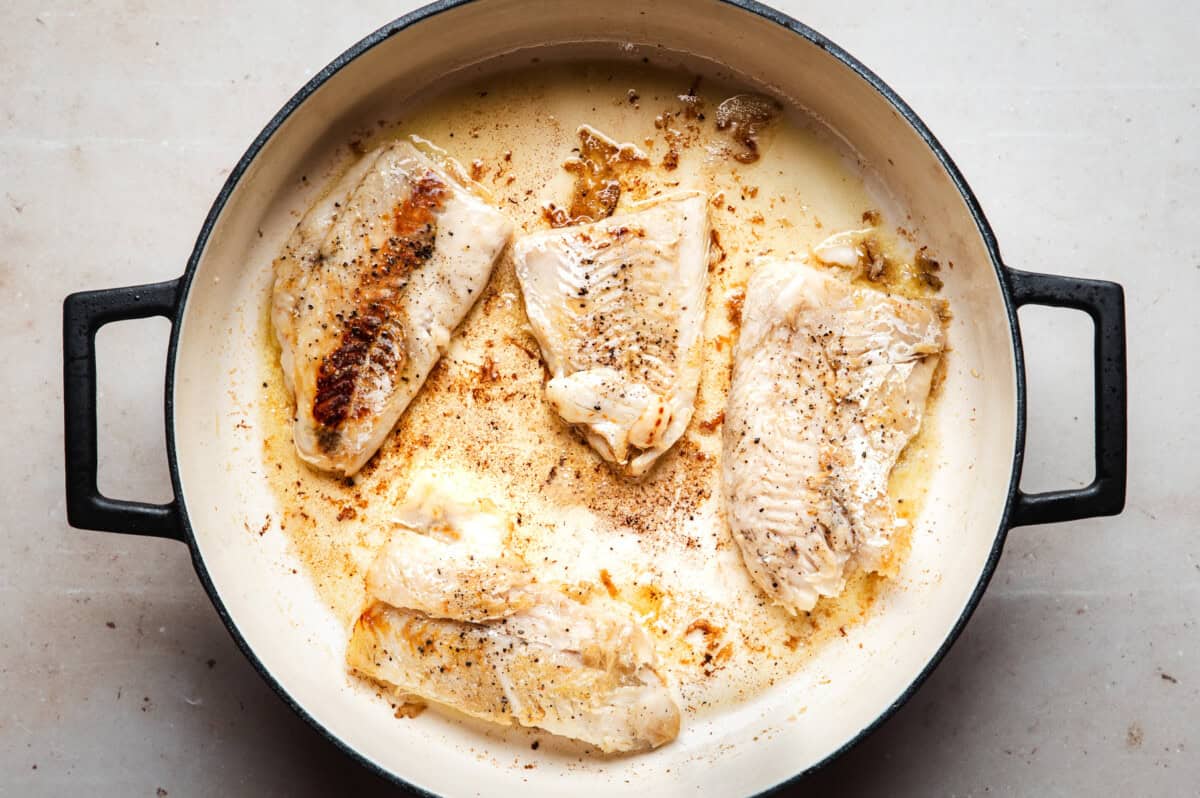 Four pieces of cod fillets are being pan-seared in a round, shallow skillet with black handles; the fillets are lightly browned and seasoned with pepper.