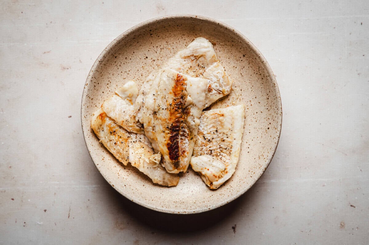 A speckled beige bowl containing several pieces of seared cod fillet, seasoned with black pepper, placed on a light-colored surface.