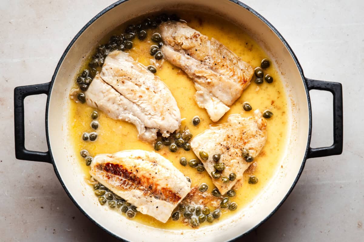 Four pieces of cooked white fish in a creamy yellow butter sauce with capers in a round, black-handled pan on a light surface.