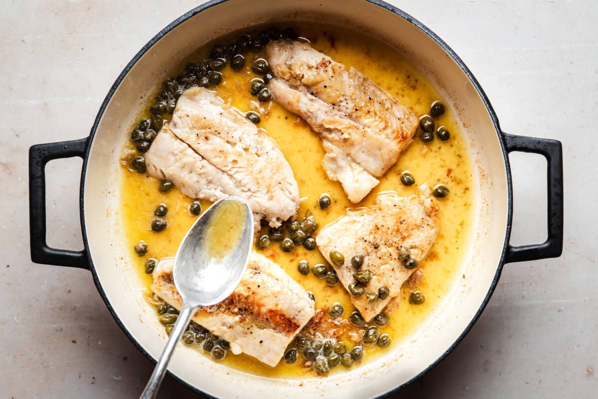 Four pieces of cooked cod fillets with capers in a lemon-butter sauce sit in a round pan, with a spoon resting inside. The pan is on a light-colored surface.
