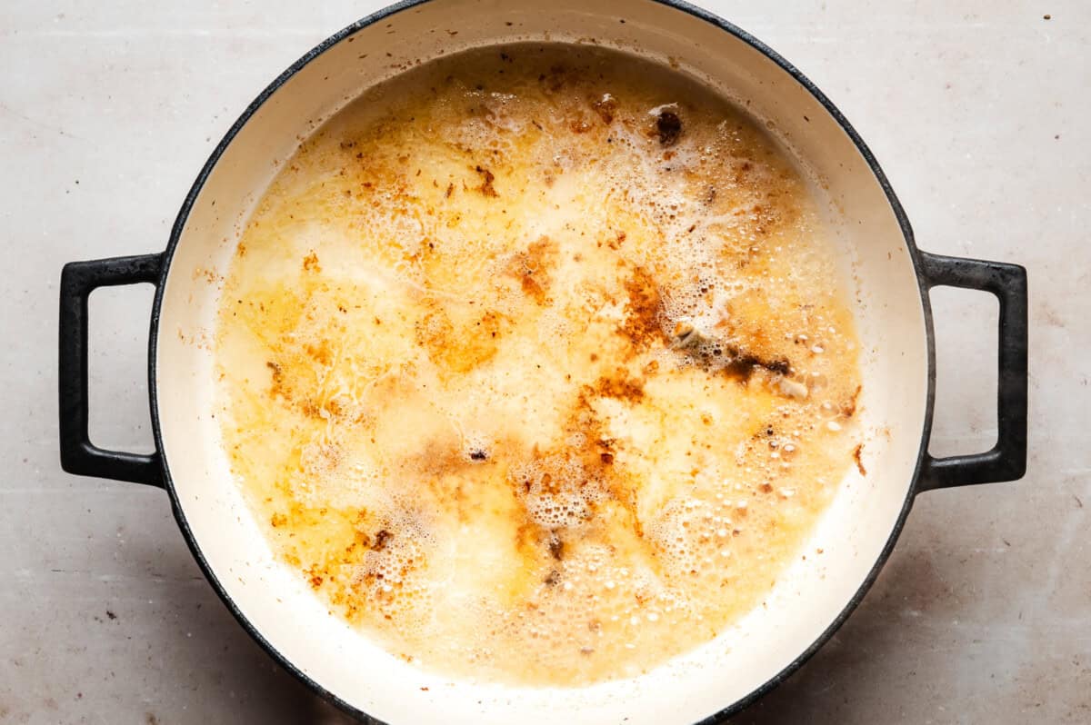 A large white pot with black handles filled with a bubbling butter and brown specks on the surface, sitting on a light-colored countertop.