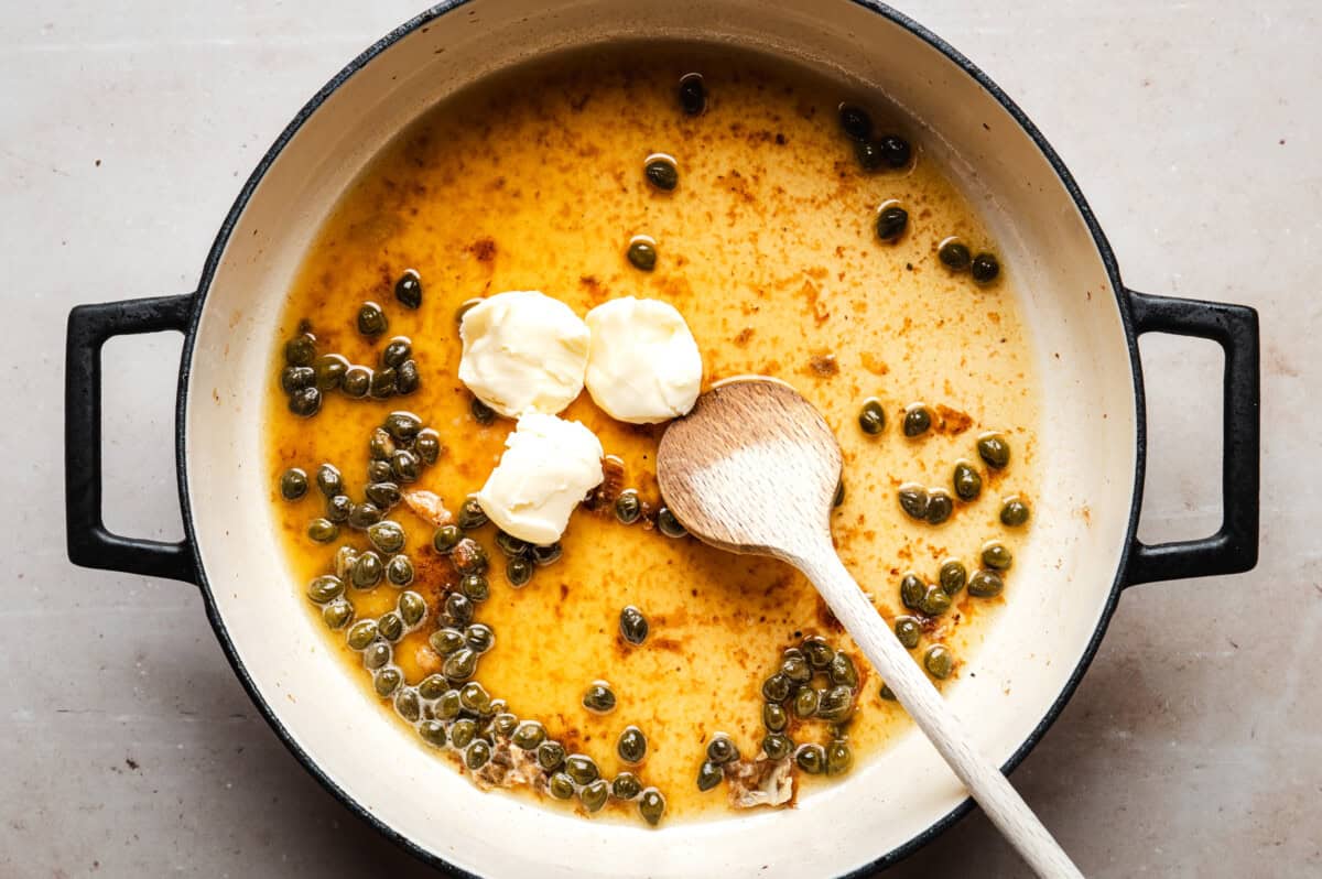 A white enameled pan contains a sauce with capers, melted butter, and a few chunks of unmelted butter. A wooden spoon rests inside the pan, stirring the mixture.