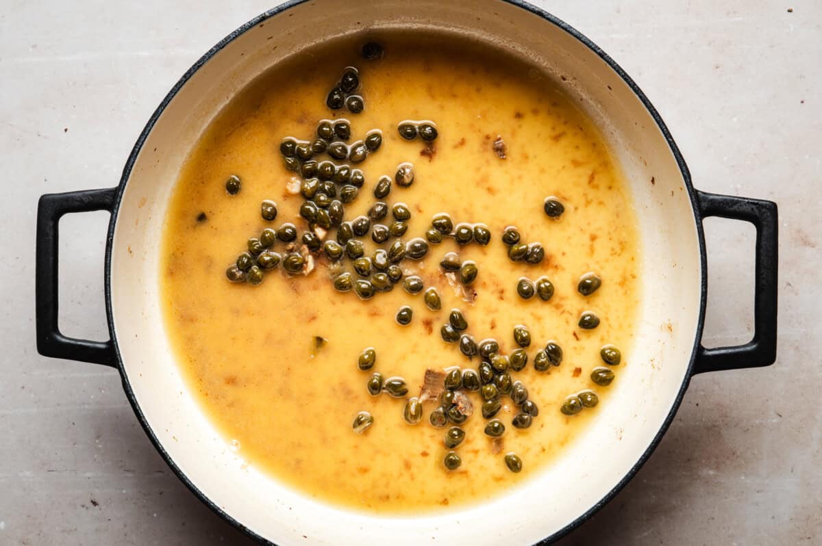 A white enameled pot with piccata sauce and capers scattered throughout. The pan is set on a light-colored surface.