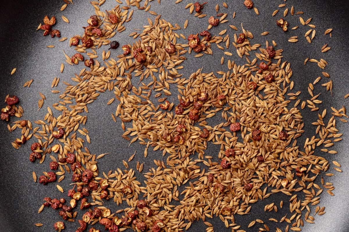 Cumin seeds and dried red Sichuan peppercorns scattered in a non-stick black pan, likely being toasted for cooking.