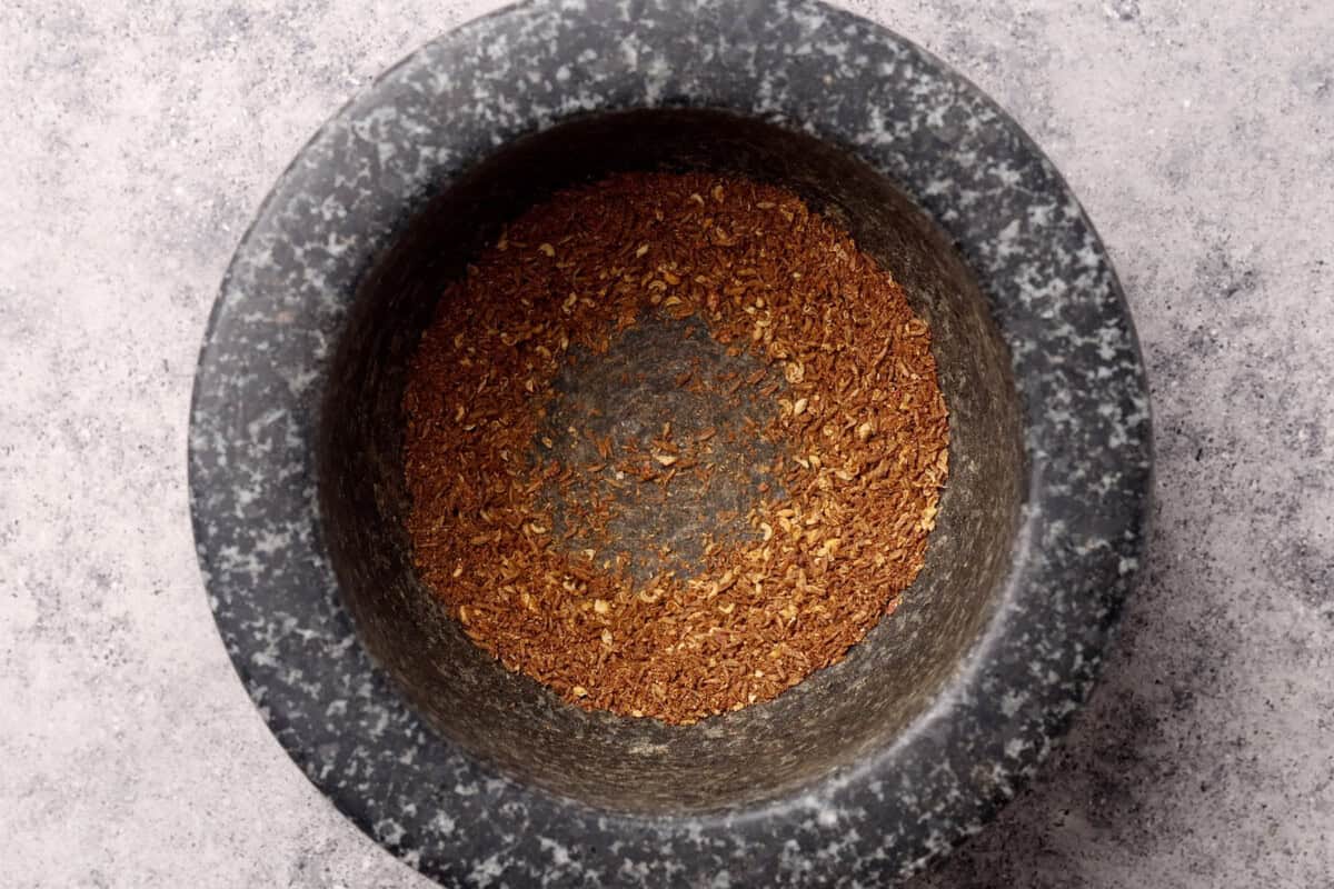 A granite mortar containing a small amount of finely ground brown spices sits on a light gray textured surface.