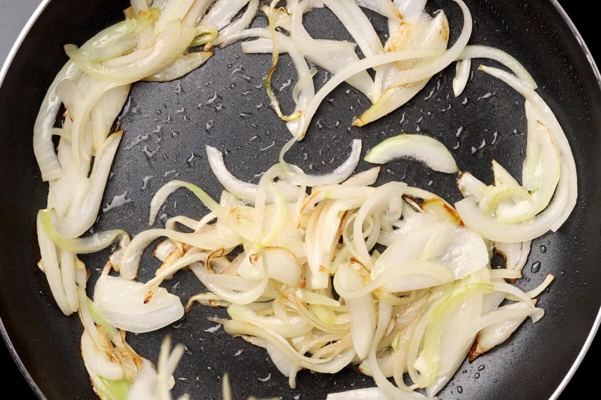 Thinly sliced onions are being saut&eacute;ed in a black frying pan. The onions are beginning to turn translucent and lightly brown, indicating they are just starting to caramelize.