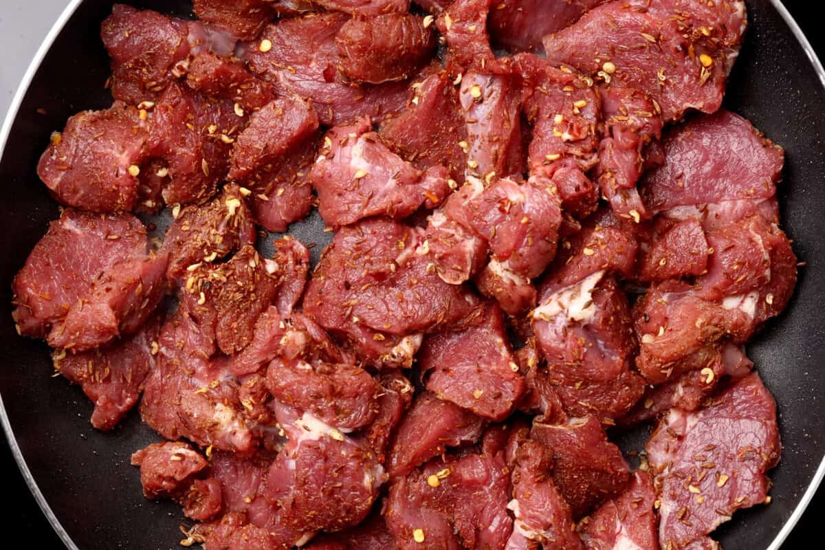 Raw marinated beef pieces with visible spices and chili flakes in a black pan, ready for cooking.