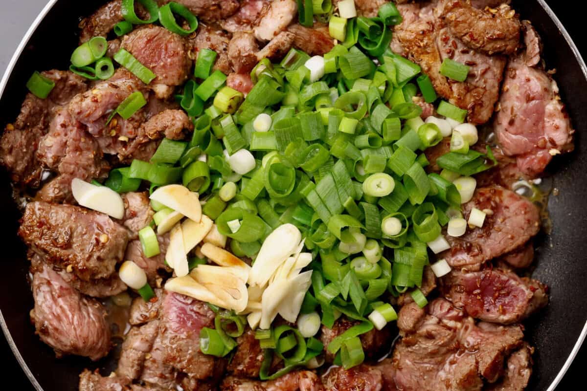Slices of browned beef are cooked in a pan with chopped green onions and thin slices of garlic, creating a colorful and appetizing stir-fry dish.