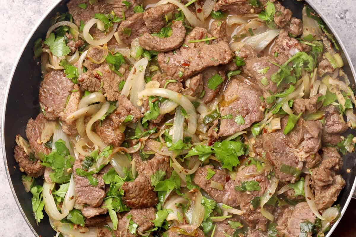 Sliced beef cooked with onions and herbs, garnished with fresh cilantro, in a pan. The dish appears savory with visible seasonings and a mix of tender meat and onion slices.
