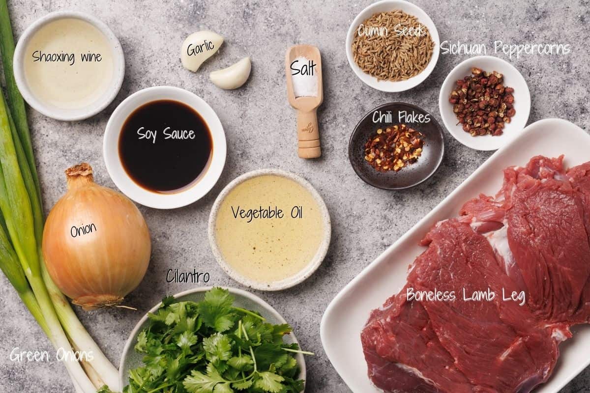 Ingredients for a cumin lamb recipe are arranged on a countertop, including a boneless lamb leg, soy sauce, Shaoxing wine, vegetable oil, garlic, onion, green onions, cilantro, salt, cumin seeds, Sichuan peppercorns, and chili flakes.