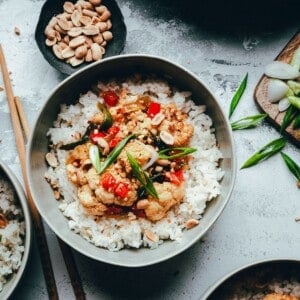 A bowl of white rice topped with kung pao cauliflower, including cauliflower and red bell pepper, garnished with sliced green onions and peanuts. Chopped peanuts and green onions are on small plates nearby. Chopsticks rest on the bowl.