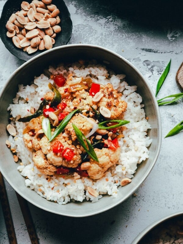 A bowl of white rice topped with kung pao cauliflower, including cauliflower and red bell pepper, garnished with sliced green onions and peanuts. Chopped peanuts and green onions are on small plates nearby. Chopsticks rest on the bowl.
