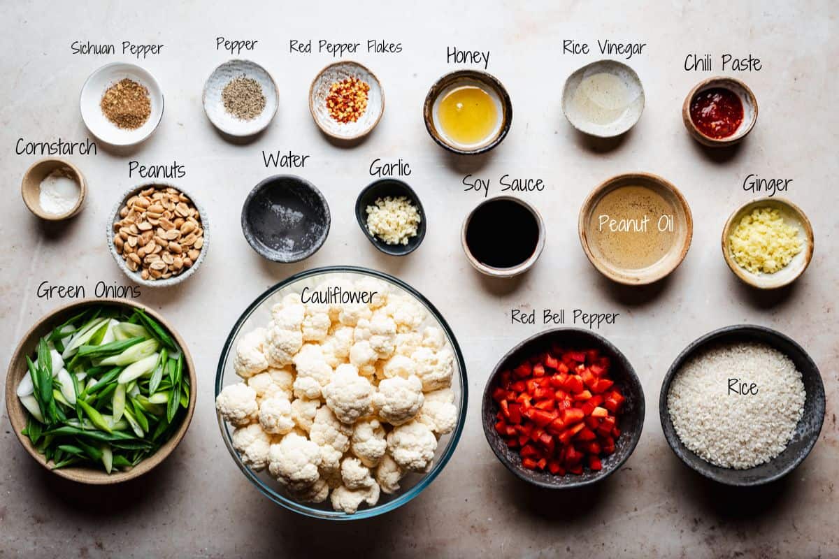 Kung Pao Cauliflower ingredients in bowls on a countertop, including chopped green onions, cauliflower florets, diced red bell pepper, rice, ginger, chili paste, peanut oil, soy sauce, garlic, water, peanuts, cornstarch, and seasonings.