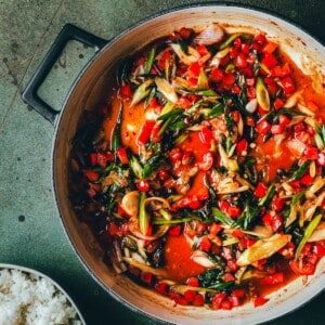 A pot filled with kung pao sauce featuring chopped red peppers, green onions, and a rich red sauce. A bowl of white rice sits partially visible beside the pot on a green surface.