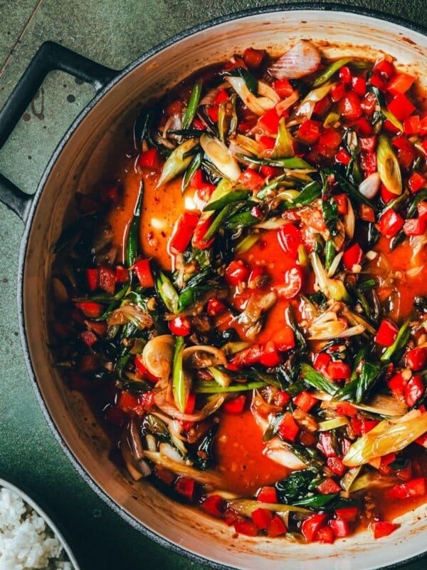 A pot filled with kung pao sauce featuring chopped red peppers, green onions, and a rich red sauce. A bowl of white rice sits partially visible beside the pot on a green surface.