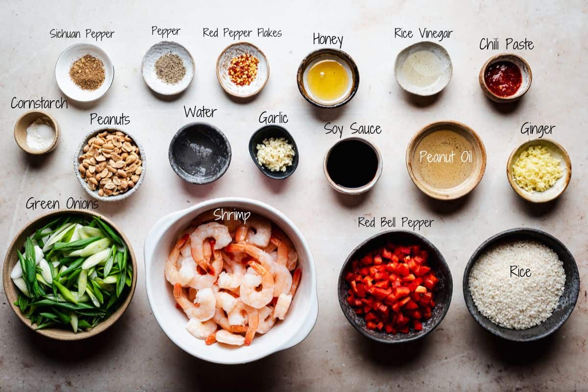 A flat lay of measured ingredients in bowls for a kung pao shrimp, including shrimp, green onions, red bell pepper, rice, peanuts, ginger, garlic, honey, soy sauce, chili paste, pepper, rice vinegar, and seasonings.