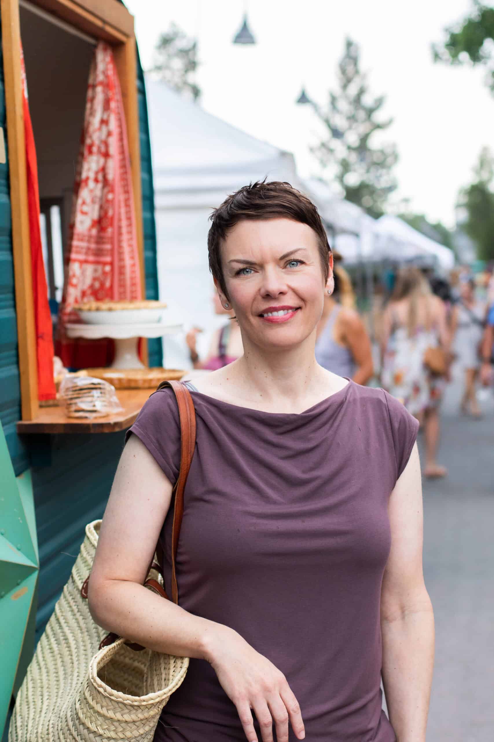 A woman with short brown hair and a purple top stands outdoors by a food truck or market stall, holding a woven bag. Tents, people, and trees line the background on a bright day.