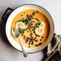 A pot of piccata sauce garnished with lemon slices and parsley, with a spoon resting inside. A green cloth sits nearby on a light-colored surface.
