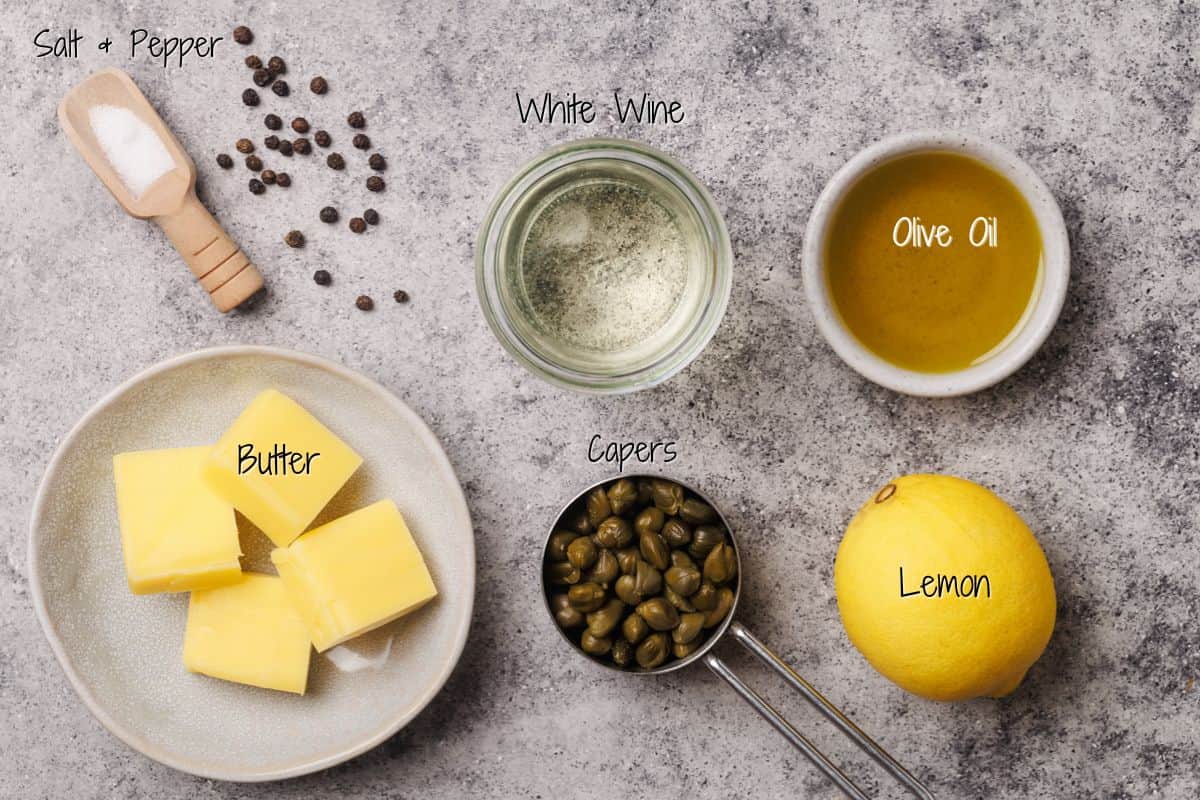 Piccata Sauce ingredients laid out on a gray surface: salt and pepper, a glass of white wine, a bowl of olive oil, a plate with cubes of butter, a measuring cup of capers, and a whole lemon. Each item is labeled.
