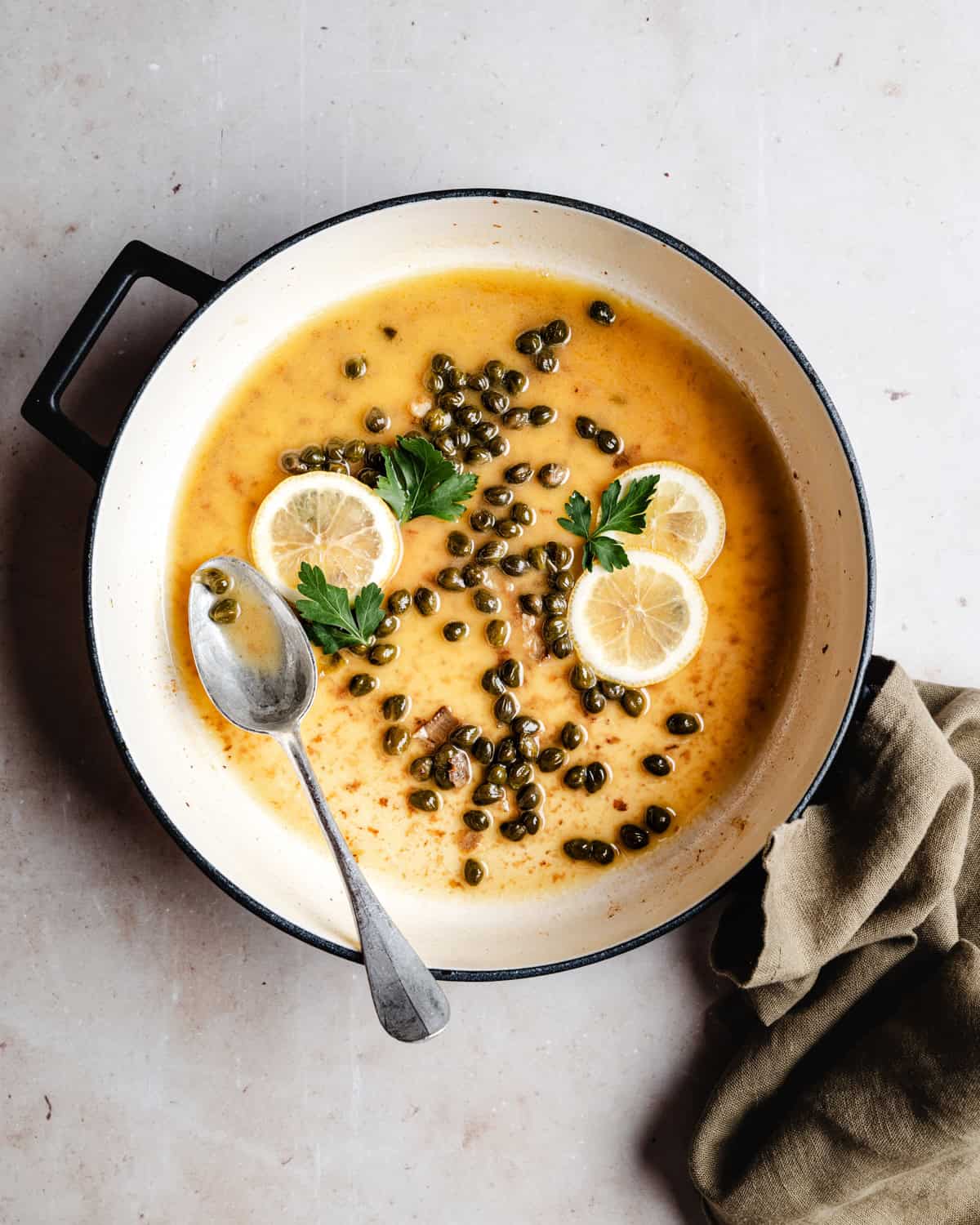 A pot filled with a Piccata Sauce, garnished with lemon slices, capers, and parsley. A metal spoon rests inside the pot, and a green cloth lies nearby on a light countertop.