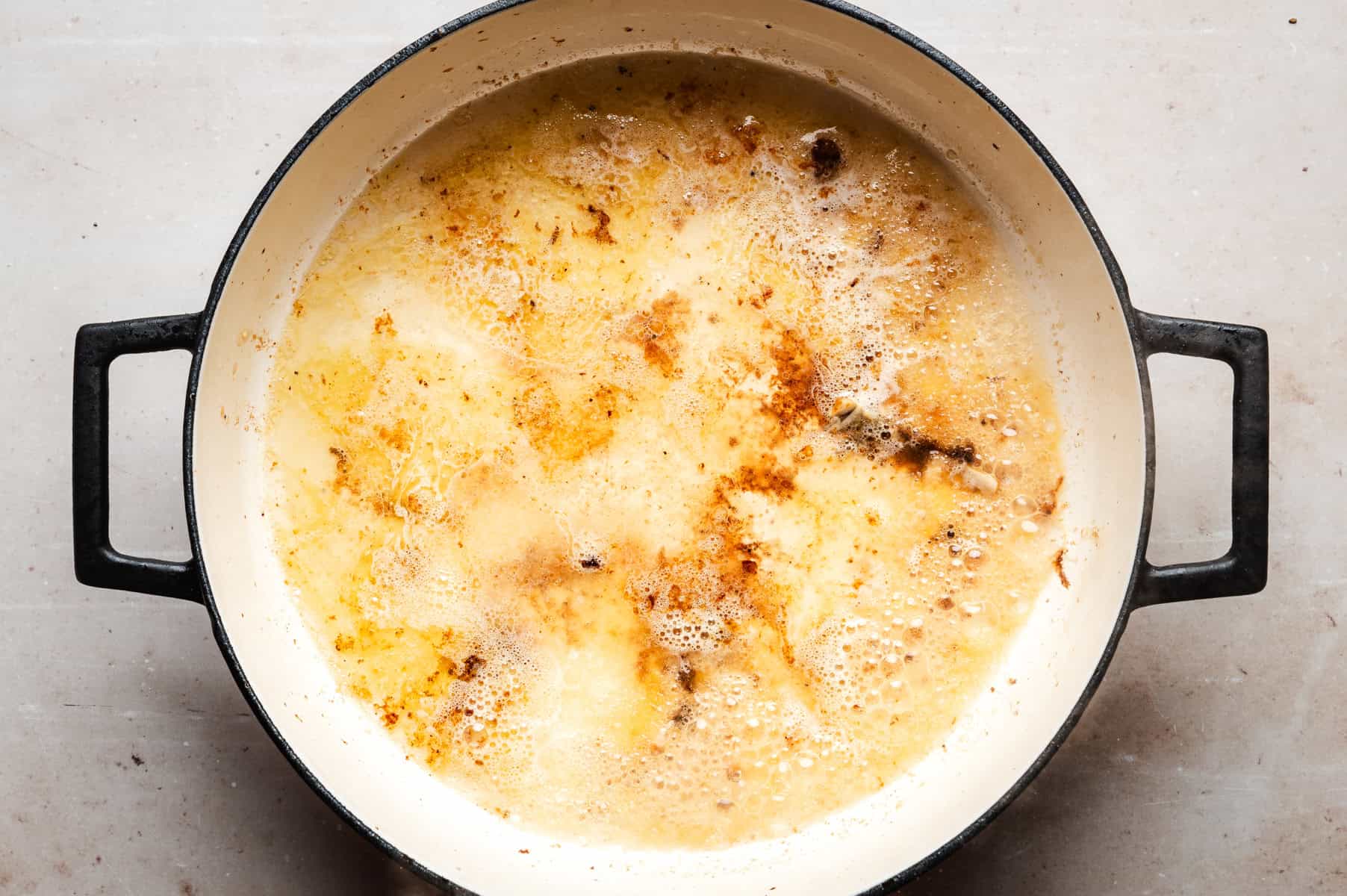 A large white pot with black handles containing a melted butter with visible bubbles and brown patches on the surface. The pot is set on a light-colored countertop.