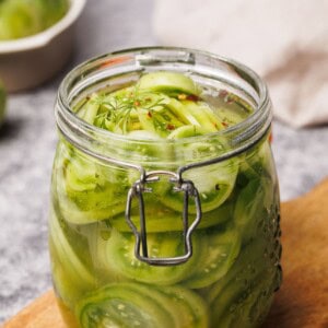 A glass jar filled with sliced green tomatoes in brine, garnished with dill, sits on a wooden surface with a blurred background.