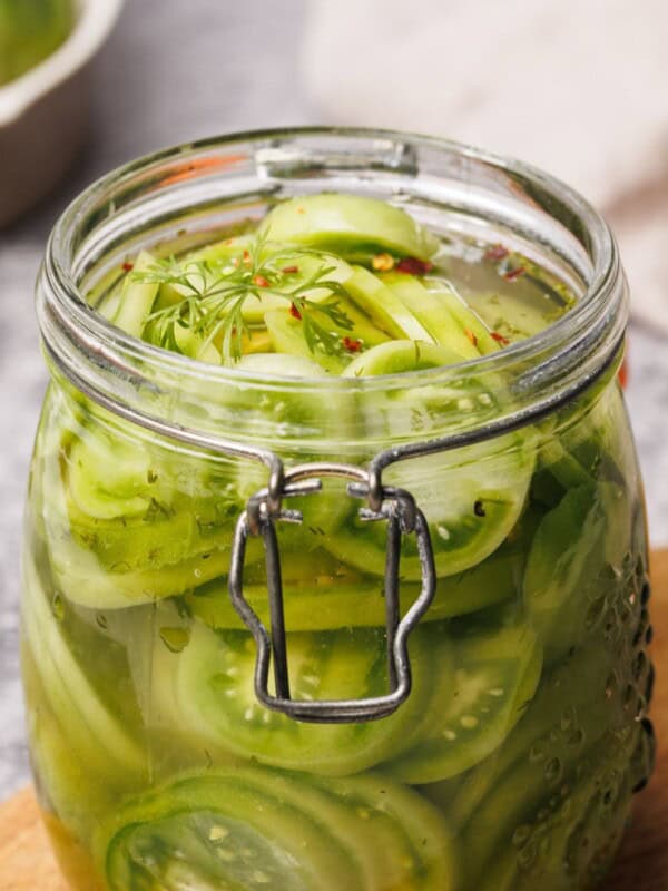 A glass jar filled with sliced green tomatoes in brine, garnished with dill, sits on a wooden surface with a blurred background.