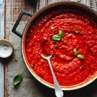 A pot of vibrant red pomodoro sauce garnished with fresh basil leaves sits on a rustic wooden table, with a spoon resting inside the sauce.
