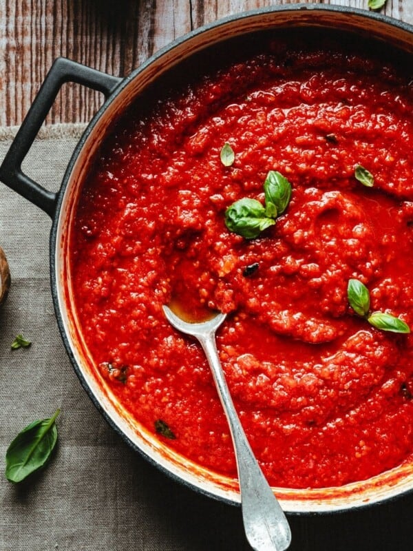 A pot of vibrant red pomodoro sauce garnished with fresh basil leaves sits on a rustic wooden table, with a spoon resting inside the sauce.
