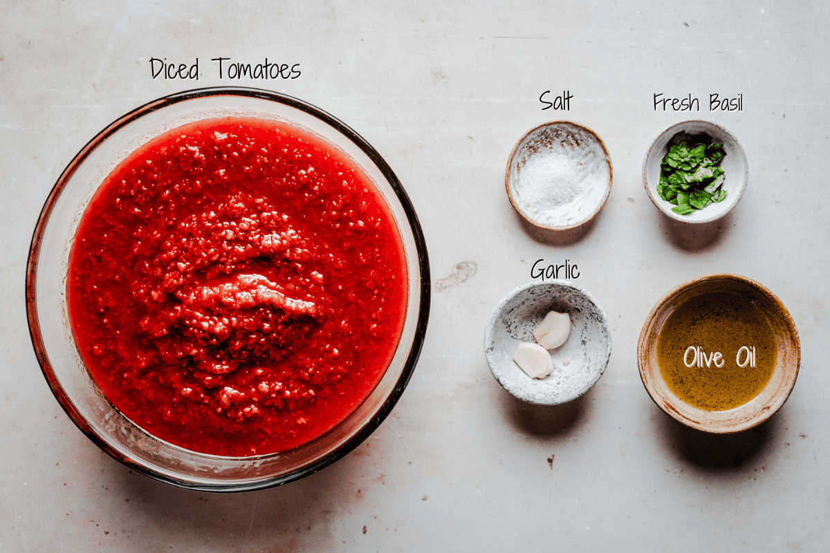 A bowl of diced tomatoes is surrounded by small bowls of salt, chopped fresh basil, two garlic cloves, and olive oil, all labeled on a light-colored surface.