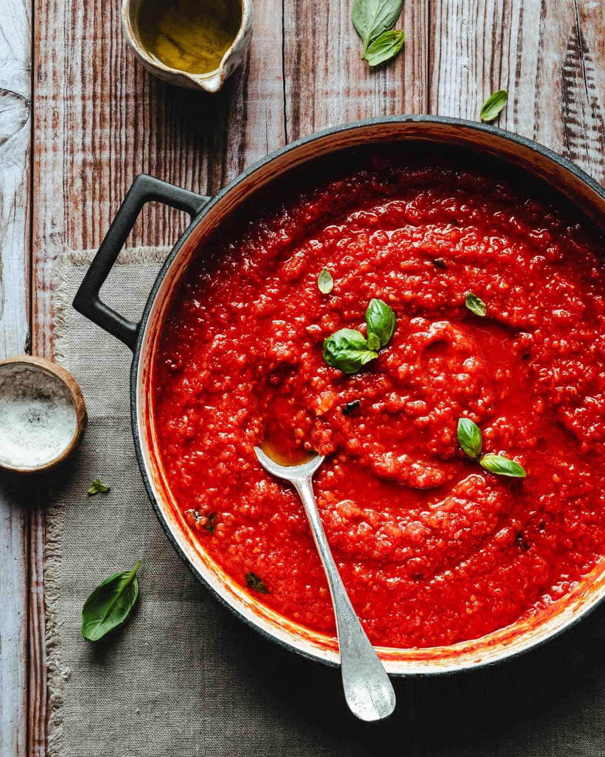 A pot of vibrant red pomodoro sauce garnished with fresh basil leaves, with a spoon resting inside, sits on a rustic wooden table alongside a small bowl of salt and a jug of olive oil.