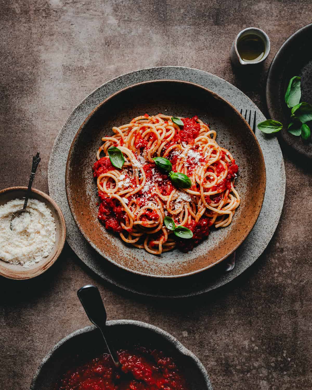 A bowl of spaghetti topped with pomodoro sauce, grated cheese, and fresh basil sits on a plate, surrounded by a small bowl of cheese, a dish of tomato sauce, a few basil leaves, and a small container of olive oil.